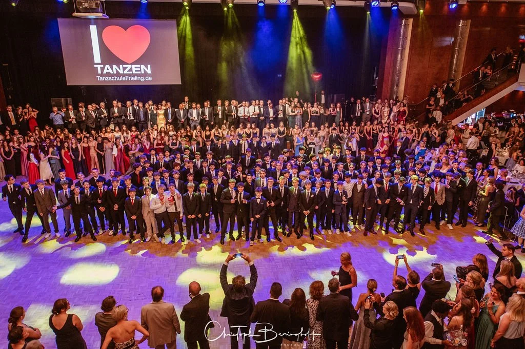 Eine große Gruppe junger Menschen in formeller Kleidung auf einer Tanzveranstaltung, im Vordergrund mehrere Personen, die Fotos machen. Im Hintergrund eine Leinwand mit einem Love-Logo und dem Slogan  "Ich liebe Tanzen", sowie bunte Bühnenbeleuchtung