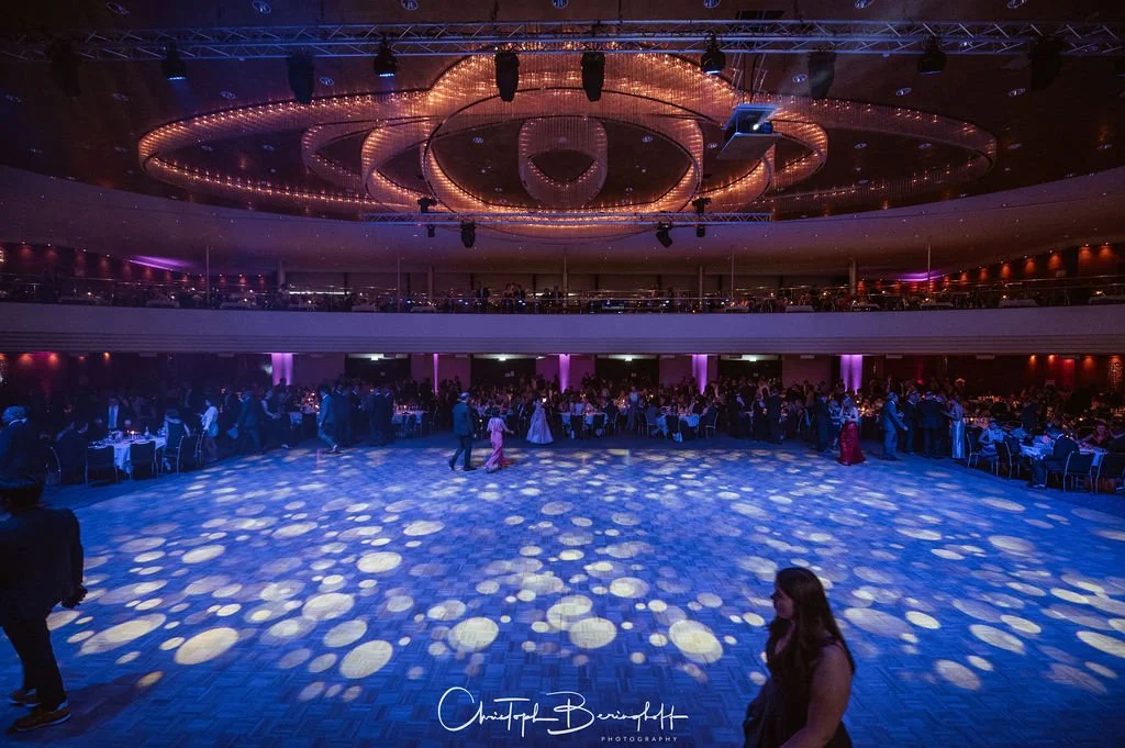 Großes Ballsaal mit tanzendem Publikum, bunte Beleuchtung, goldene Kronleuchter an der Decke, Tanzfläche in der Mitte, Sitzgelegenheiten am Rand, elegante Abendkleidung.