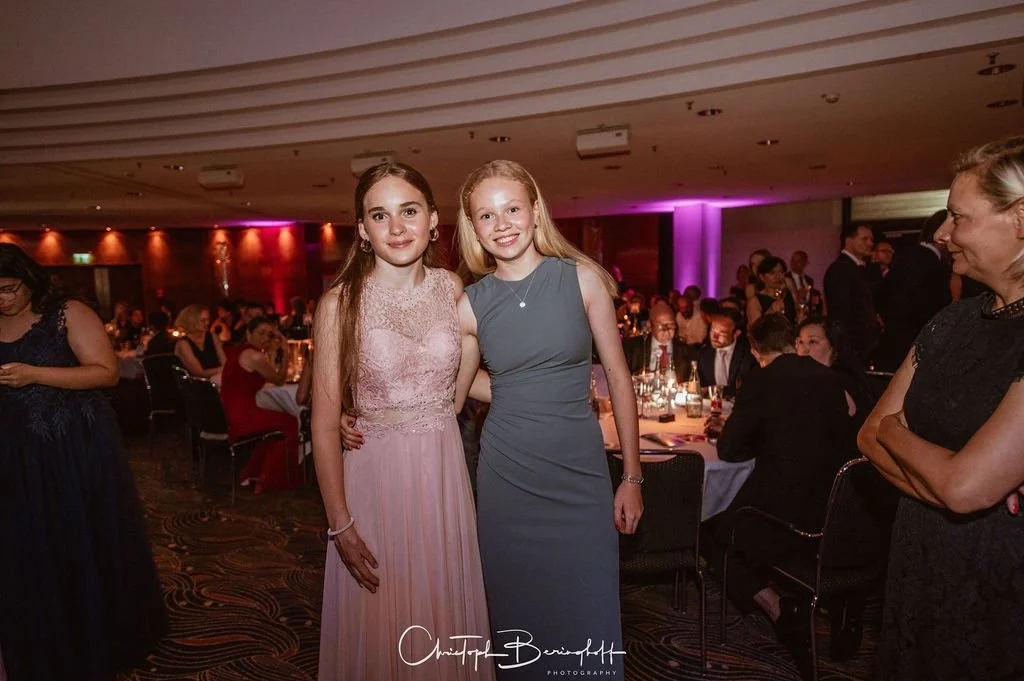 Zwei junge Frauen in formeller Abendkleidung bei einer Gala-Veranstaltung, umgeben von anderen Gästen im festlich dekorierten Raum.