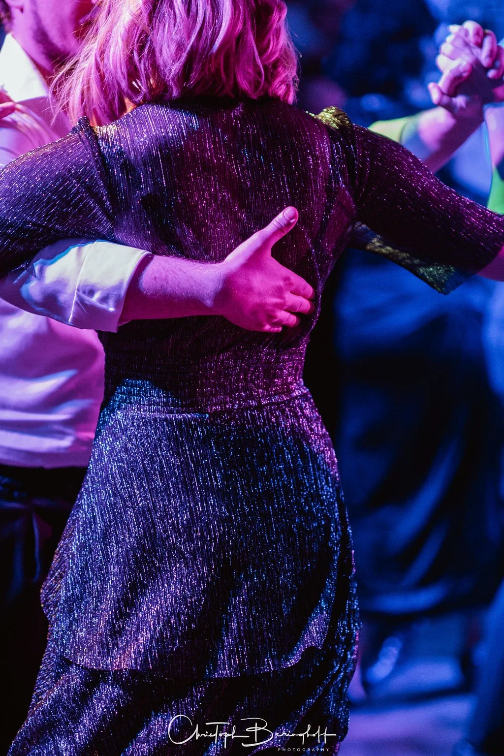 Zwei Menschen tanzen eng auf einer Party, eine Person trägt ein glänzendes schwarzes Kleid, während die andere Person eine weiße Bluse trägt. Es herrscht eine lebendige, bunte Atmosphäre.