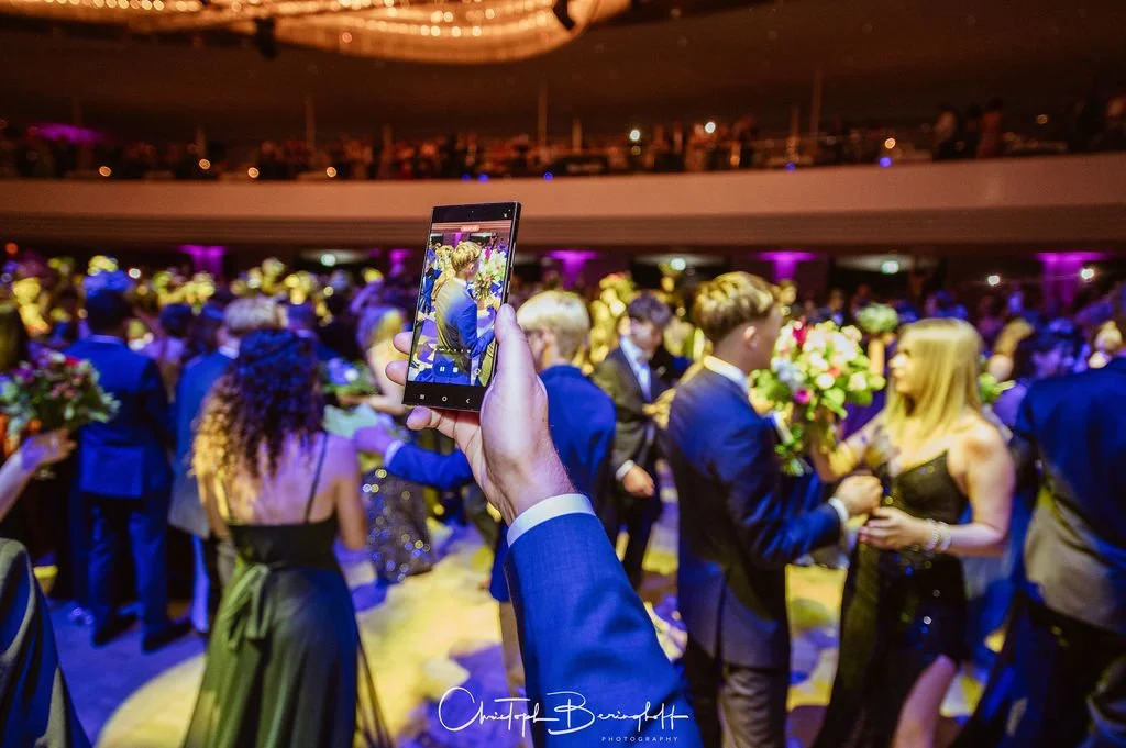 Menschen bei einer formellen Veranstaltung, die sich mit Blumen und in eleganter Kleidung befinden, während eine Person ein Smartphone benutzt, um den Moment festzuhalten.