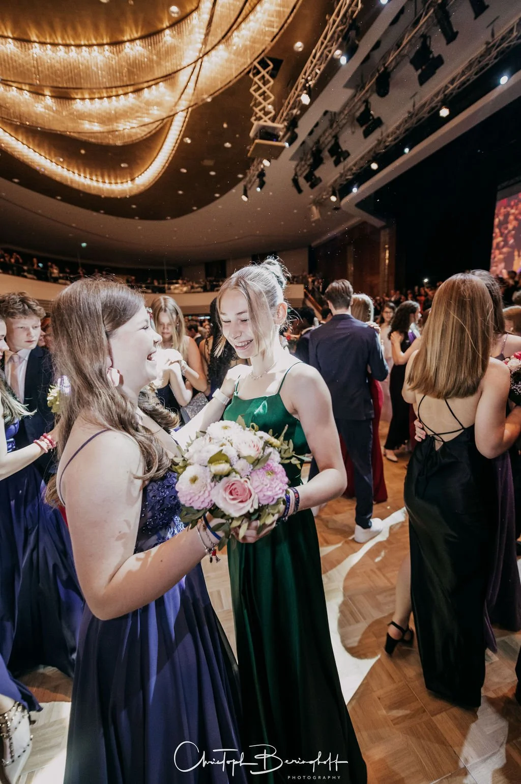 Zwei lachende junge Frauen bei einer Feier im festlichen Abendkleid, eine hält einen Blumenstrauß, im Hintergrund viele weitere Menschen in elegant gekleidet, in einem großen Veranstaltungsraum mit goldener Decke und Bühne.