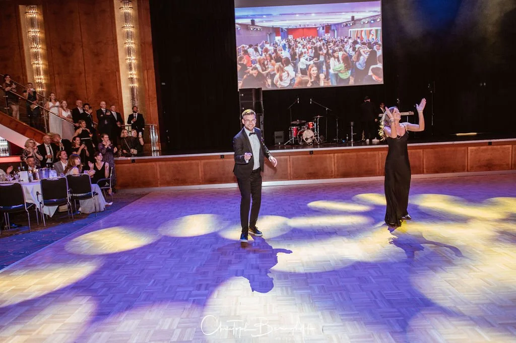 Ein Mann und eine Frau tanzen auf einer Ballenhalle mit Publikum und einer großen Leinwand im Hintergrund.