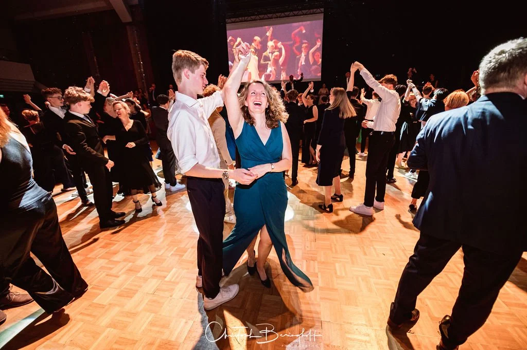 Menschen tanzen auf einer Party oder einem Ball in einem festlich dekorierten Raum, mit einer großen Leinwand im Hintergrund, auf der eine andere Tanzszene gezeigt wird.