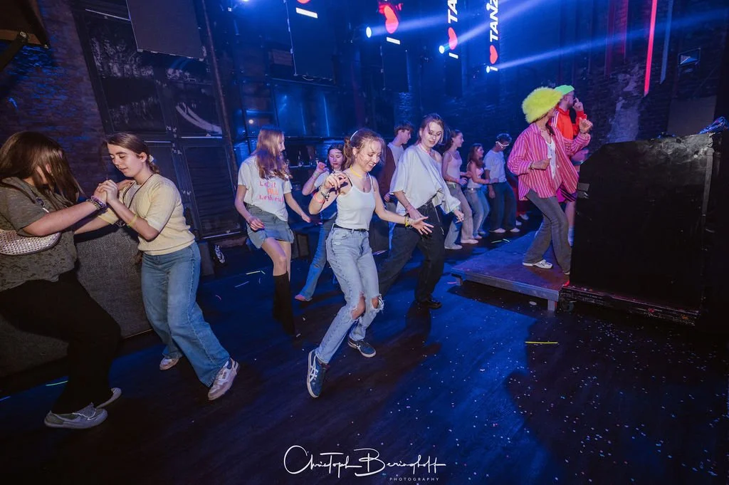 Teenagers are dancing in a nightclub, with colorful lights, a DJ booth in the background, and a cheerful atmosphere.
