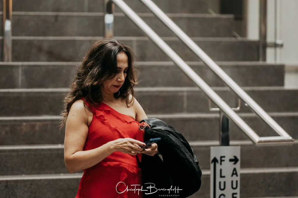 Frau mit braunen, lockigen Haaren in rotem Kleid steht an einer Treppe im U-Bahnhof und schaut auf ihr Smartphone.