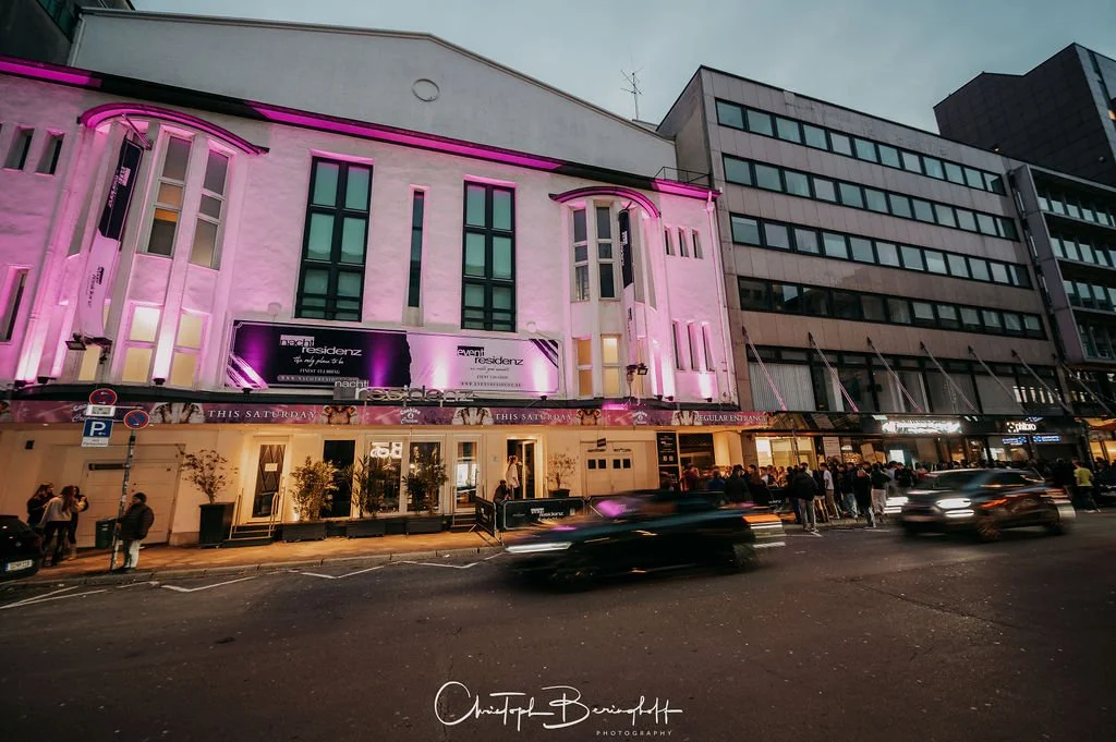 The photo shows a multi-story building illuminated by pink lights at night. People are standing on the street in front of the building, and cars are moving by. The building appears to be a club or venue, with a line of people waiting...