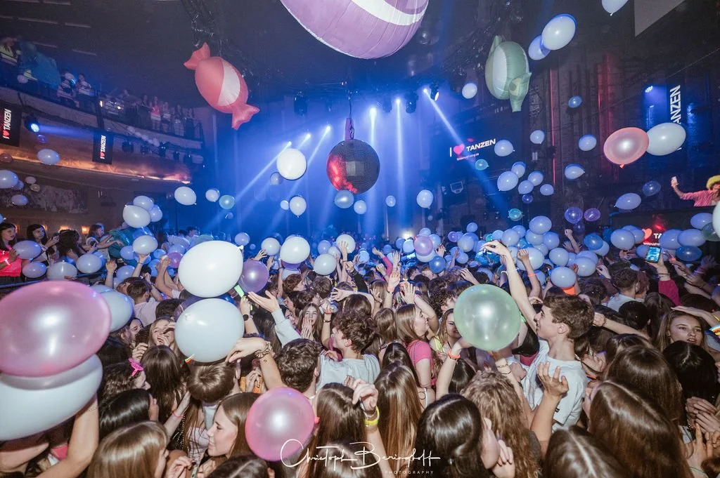 People are celebrating at a party with balloons and dancing in a dark room, colorful lights, and a large dance floor.