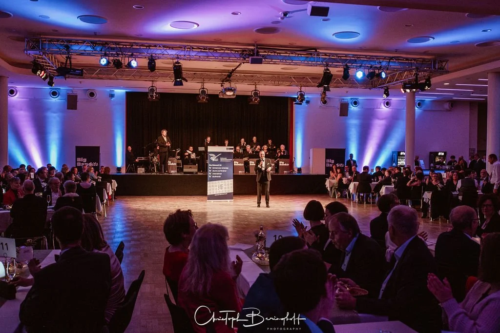 Ein festlich gestalteter Saal mit einer Bühne, auf der eine Rede gehalten wird, und einem Orchester im Hintergrund. Der Saal ist mit buntem Licht beleuchtet, und die Gäste sitzen an Tischen und applaudieren.