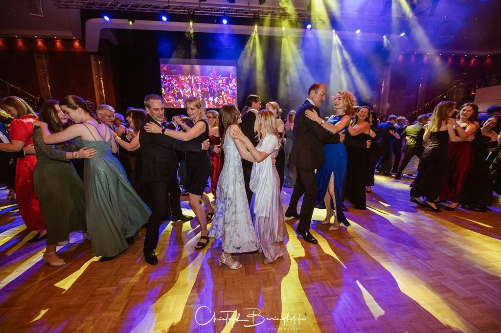 Menschen tanzen auf einer festlichen Veranstaltung im Saal, gekleidet in elegante Abendkleidung, mit buntem Licht- und Schatteneffekt auf dem Parkettboden.