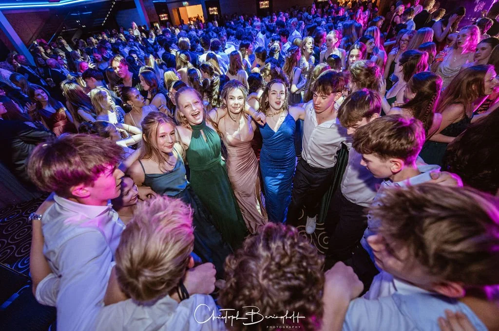 Junge Leute tanzen und feiern auf einer Party mit bunten Lichtern und einer großen Menschenmenge im Hintergrund.