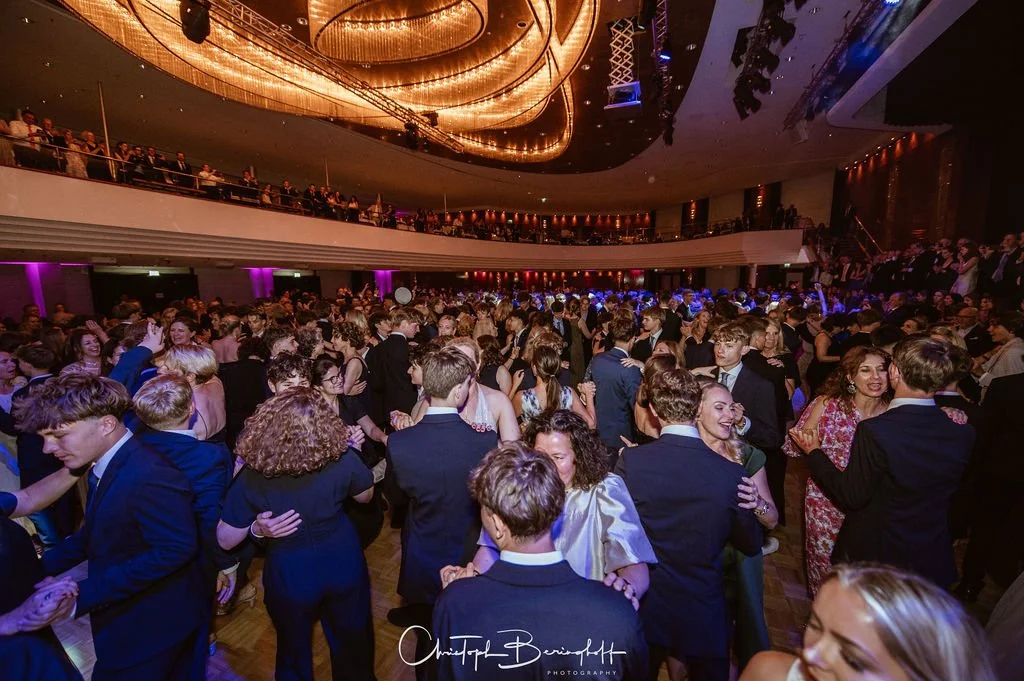 Tanzende Menschen bei einer Party in einem großen Ballsaal, mit einer Mischung aus jungen und älteren Gästen, eleganter Kleidung und bunten Lichtern.