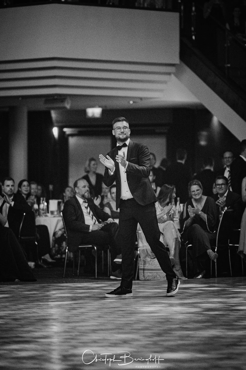 Schwarz-weiß-Foto eines Mannes im Anzug, der auf einer Tanzfläche bei einer Veranstaltung tanzt, umgeben von einem Publikum, das applaudiert und lacht.