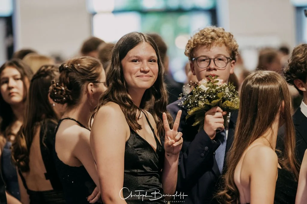 Jugendliche bei einer festlichen Veranstaltung, wobei ein Mädchen in der Mitte lächelnd die Frieden-Geste zeigt und ein Junge neben ihr ein Blumenbouquet hält.