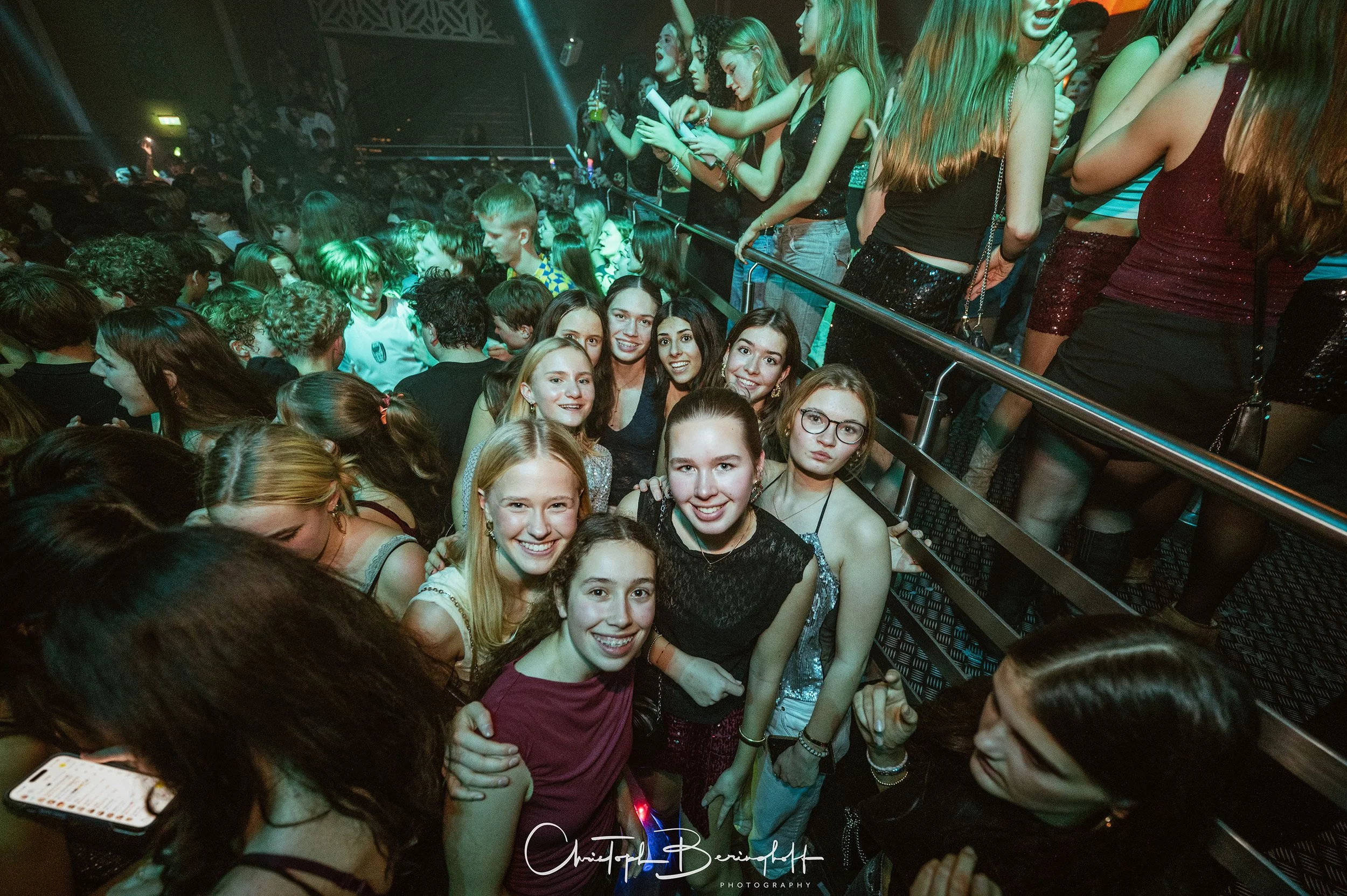 Gruppe junger Frauen in einer Partykneipe, offensichtlich beim Feiern, mit leuchtenden Lichtern und einer großen Menschenmenge im Hintergrund.