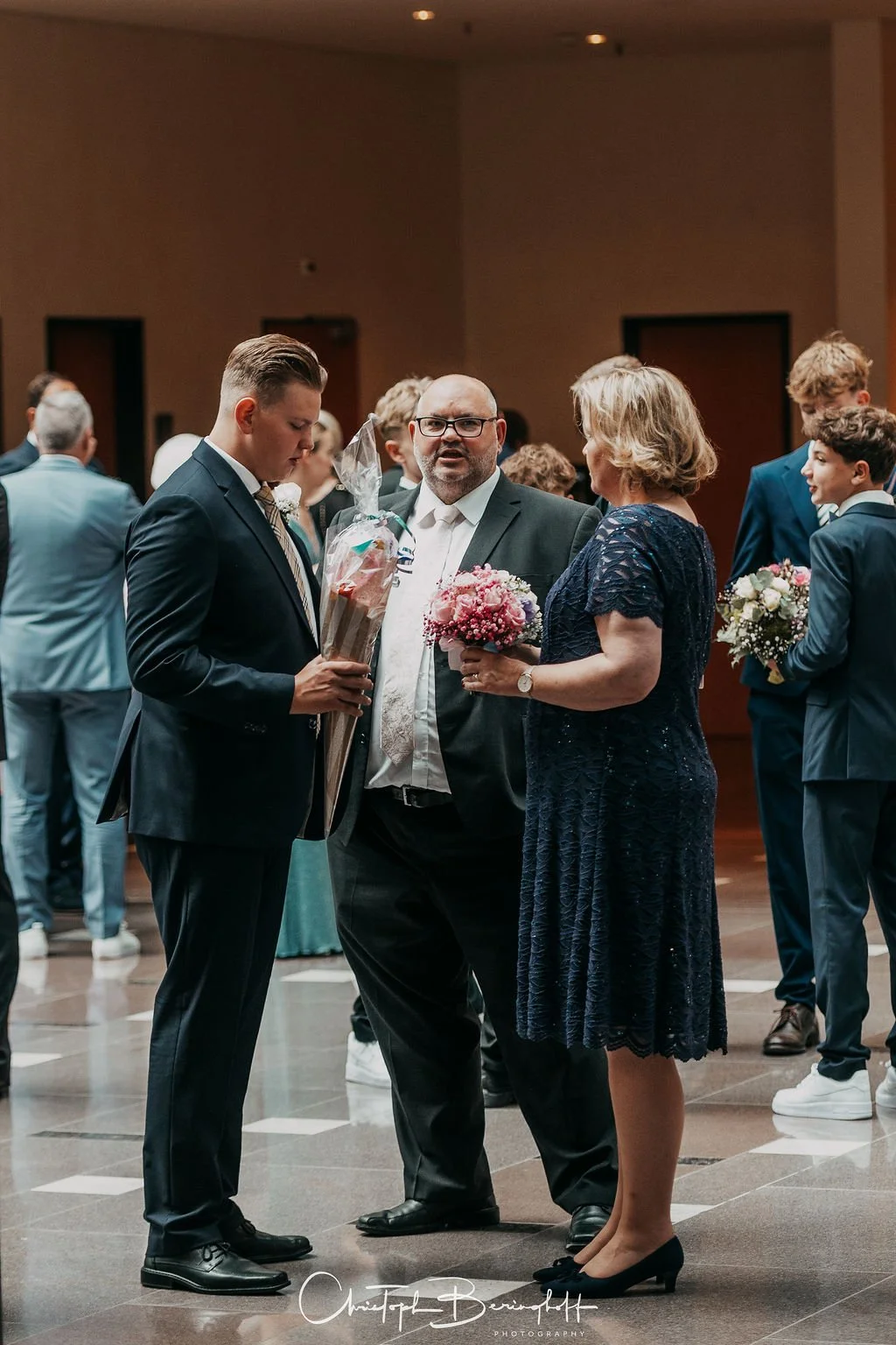 Ein Mann in Anzug hält ein Geschenk und spricht mit einer Frau in einem dunklen Kleid, während andere Gäste im Hintergrund bei einer Feier sind.