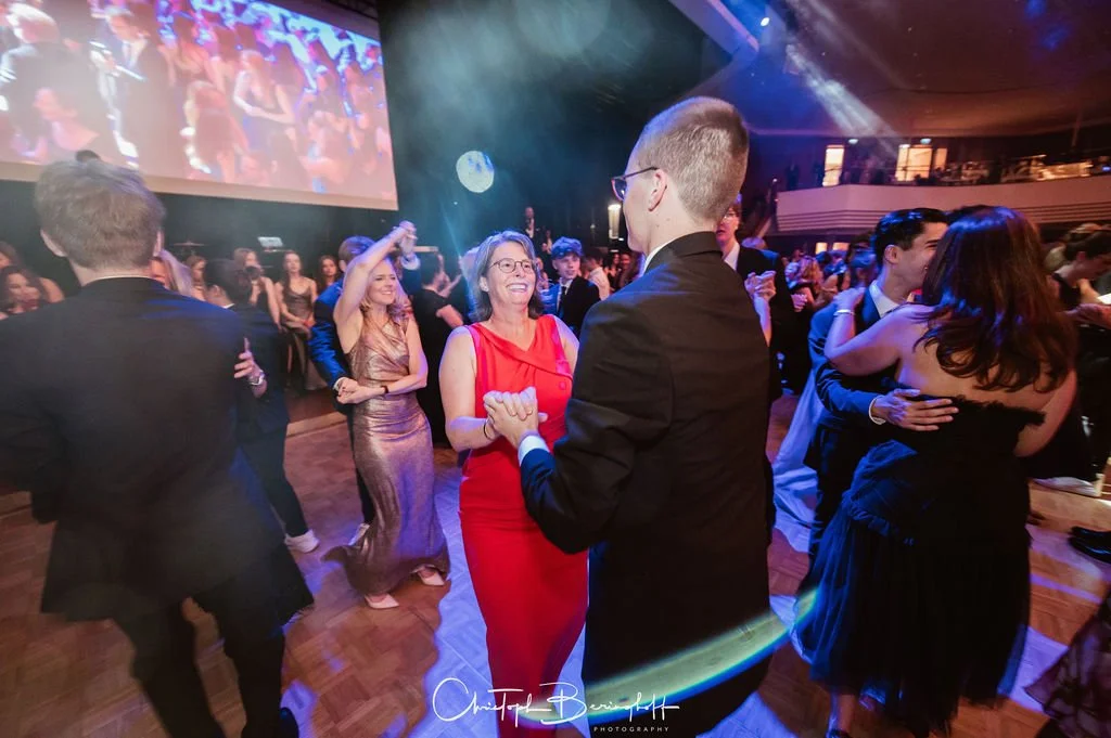 Menschen tanzen auf einer Party oder einem Ball, lachen und haben Freude, im Hintergrund eine große Leinwand mit einem Video. Es herrscht lebendige, festliche Atmosphäre.