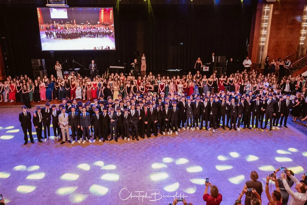 Große Gruppe von jungen Männern in Anzügen im Vordergrund und Frauen in eleganten Kleidern im Hintergrund, die auf einer festlichen Bühne bei einem Event stehen, mit einem großen Bildschirm im Hintergrund, der eine Gruppenaufnahme zeigt.