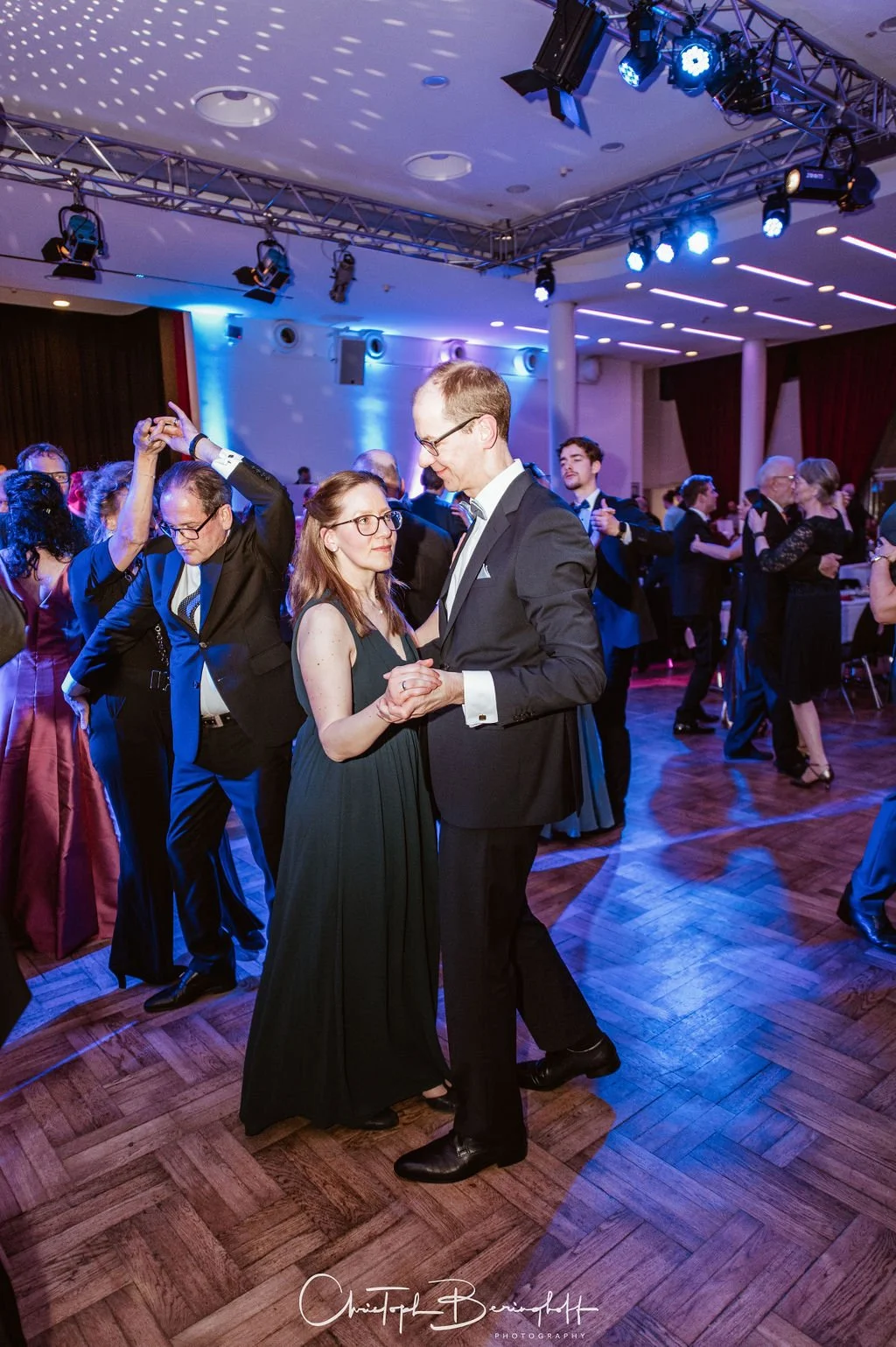 Tanzpaar tanzt auf einem festlichen Ball. Andere Paare tanzen im Hintergrund. Der Raum ist modern dekoriert mit blauen Beleuchtungen an der Wand und Decke.
