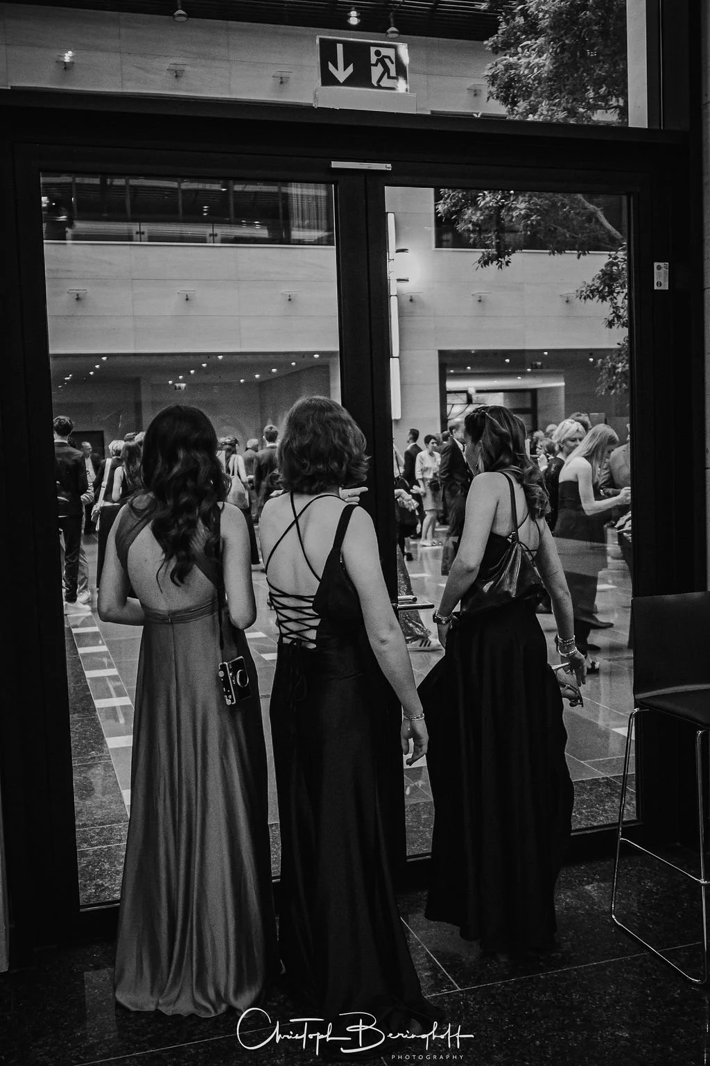 Drei Frauen in eleganten schwarzen Kleidern stehen vor einer Glastür, durch die eine Menschenmenge im inneren eines modernen Gebäudes sichtbar ist. Oben ist ein Fluchtweg-Schild sichtbar.