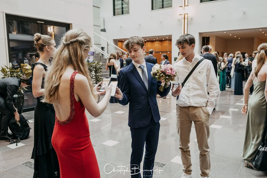 Zwei junge Männer in Anzügen und ein junge Frau in einem roten Kleid posieren für ein Foto in einer modernen, hellen Hotellobby mit anderen Gästen im Hintergrund.
