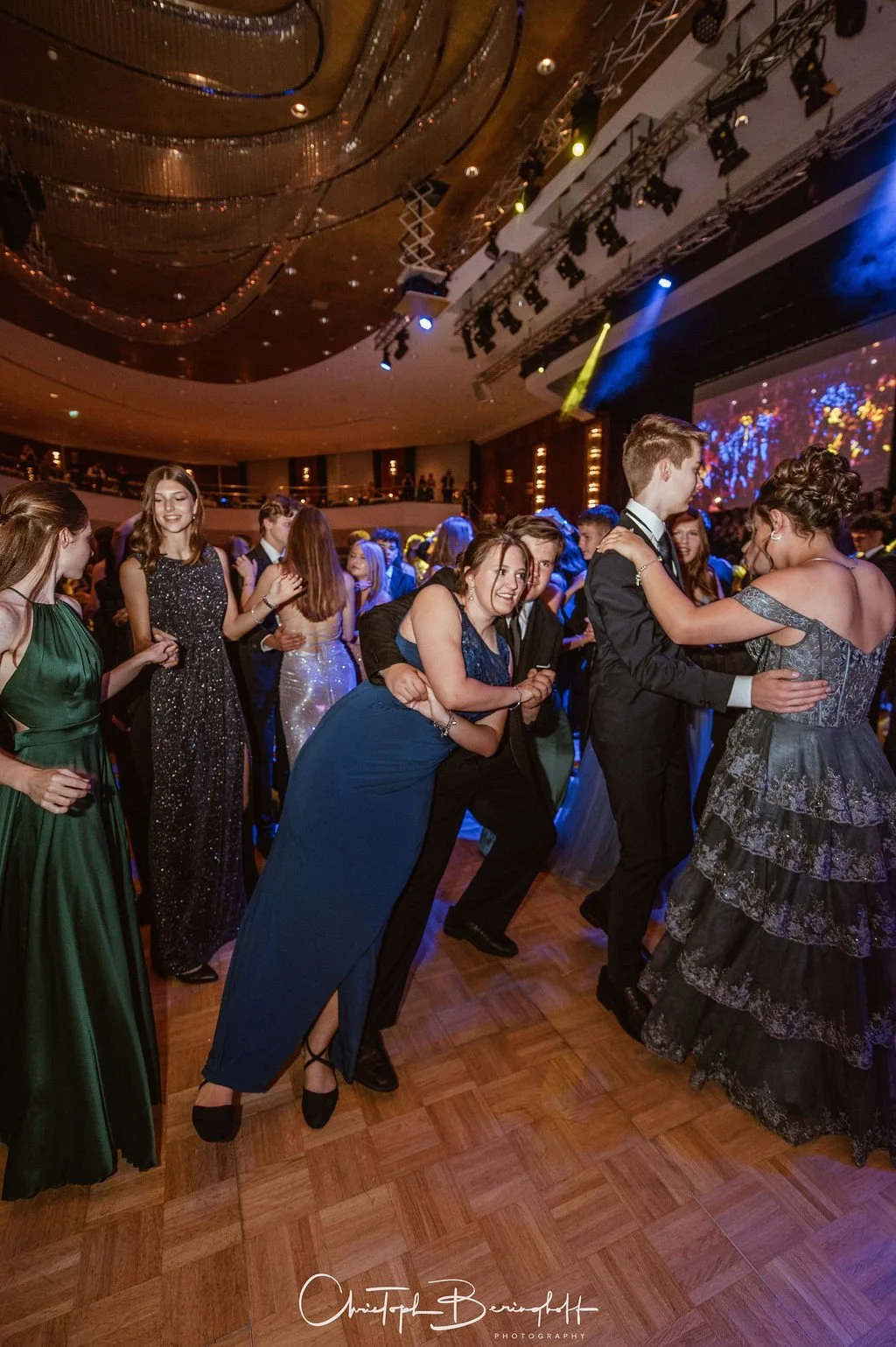 Junge Menschen tanzen bei einer formellen Veranstaltung in eleganten Kleidern und Anzügen in einem festlich dekorierten Saal mit buntem Licht und einer großen Bühne