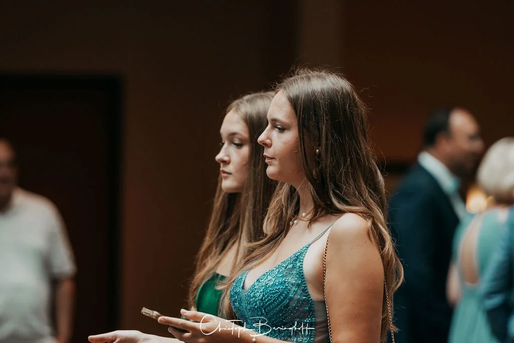 Zwei Frauen stehen nebeneinander auf einer Party, eine trägt ein türkisfarbenes Kleid und hält ein Smartphone, die andere trägt ein grünes Kleid. Im Hintergrund sind weitere Gäste zu sehen.
