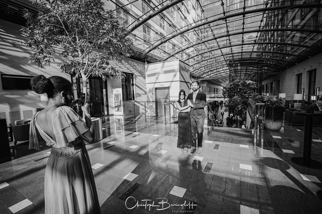 Zwei Menschen posieren für ein Foto in einem modernen, hell erleuchteten Gebäude mit Glasdecke, während eine Frau in einem eleganten Kleid sie fotografiert.