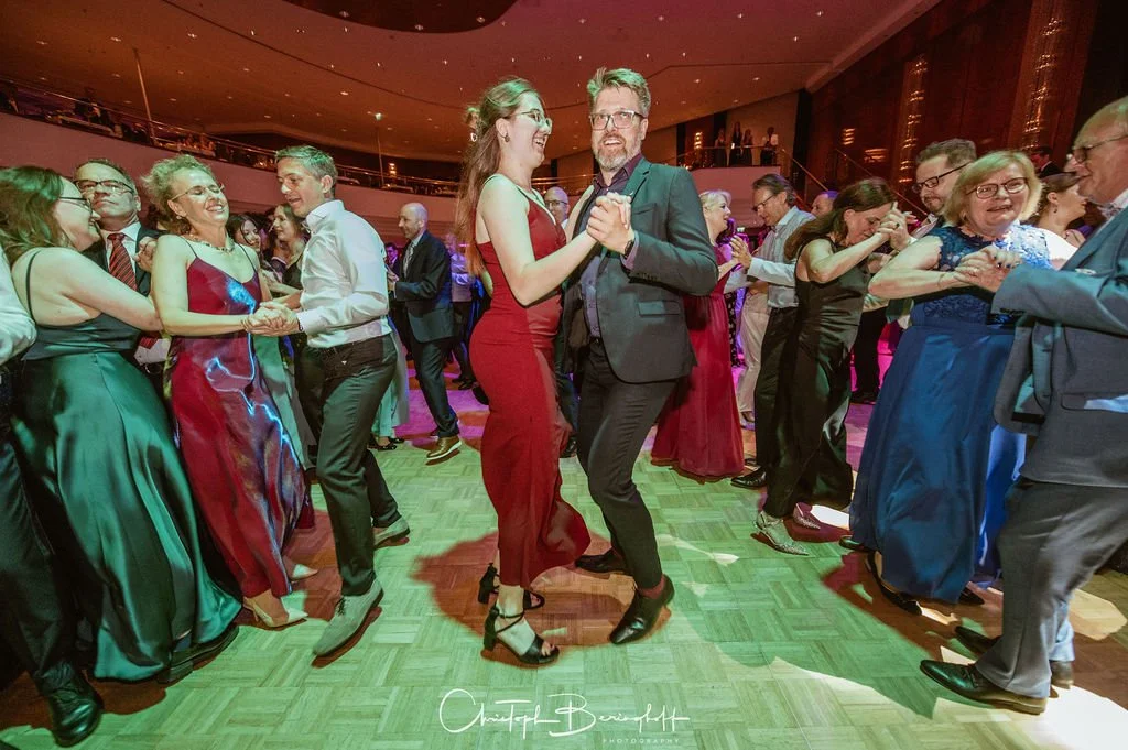 Menschen tanzen auf einem festlich geschmückten Ballsaal, viele tragen elegante Abendkleidung.