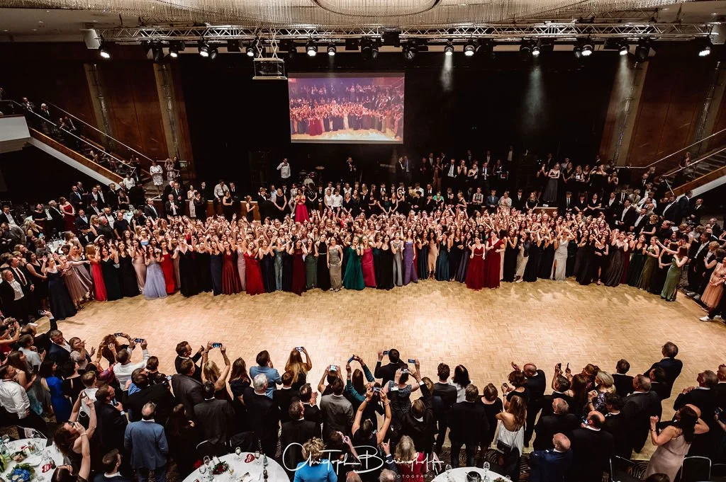 Große Abschlussball mit zahlreichen Tänzern in festlicher Kleidung auf Tanzfläche, umgeben von Gästen in eleganter Abendgarderobe, im Hintergrund Bühne mit projecting singt. Viel Beifall und Kameraaufnahmen. Moderner Festsaal mit Metalltrennwänden.