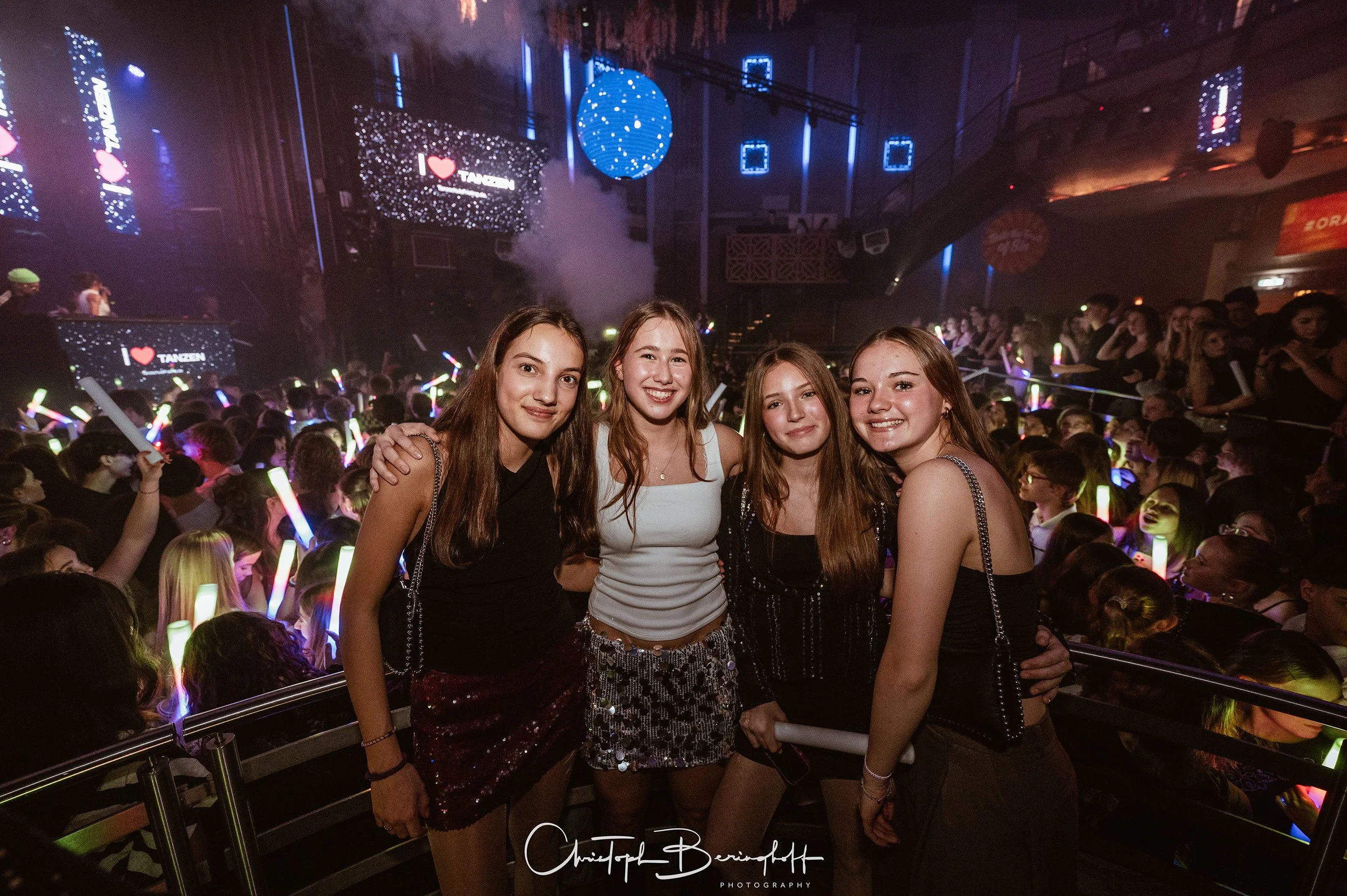 Vier junge Frauen posieren im Tanzclub, umgeben von einer großen Menschenmenge, die mit Lichtstäben tanzt, mit Neonlichtern und einem DJ-Bereich im Hintergrund.
