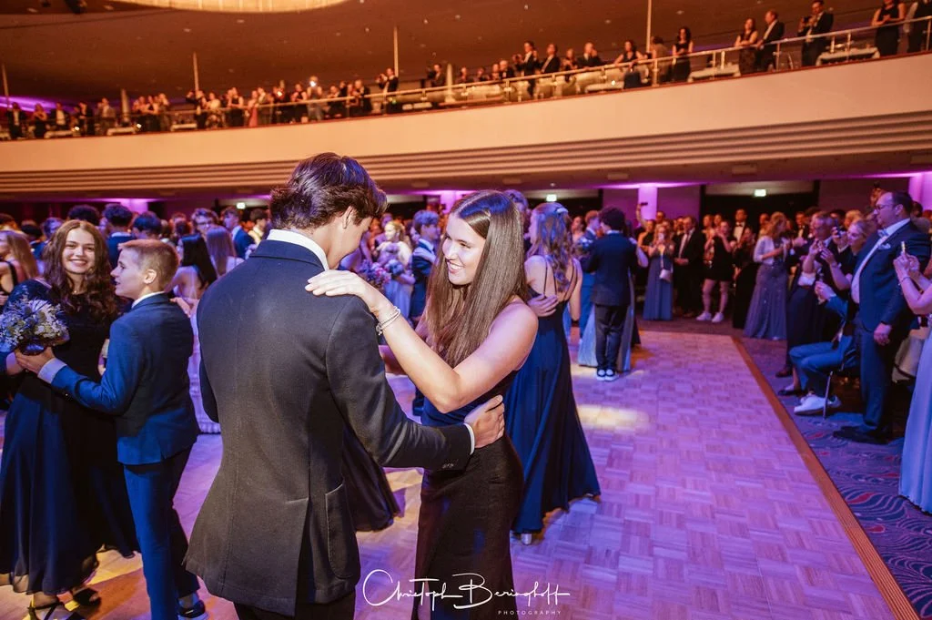 Tanzen auf einem festlichen Ball, junge Paare in eleganter Kleidung, im Hintergrund weitere Gäste und eine Mehrgeschoss-Architektur mit Balustrade, pinke und blaue Beleuchtung.