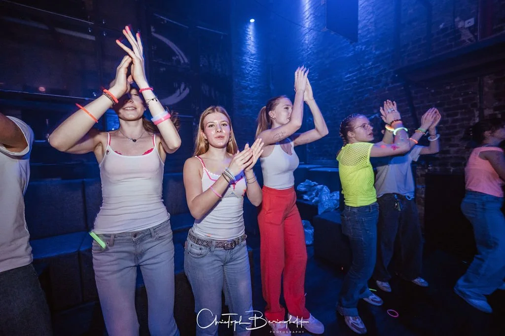 People are dancing in a club setting with dark brick walls, neon lights, and several people expressing rhythm and joy.