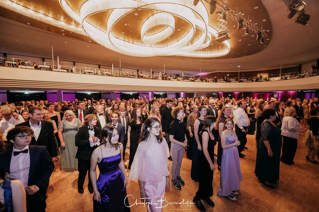 Menschen bei einer formellen Veranstaltung in einem großen Saal, viele tragen elegante Kleidung wie Abendkleider und Anzüge.
