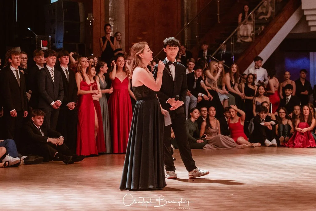 Junge Männer und Frauen in formeller Kleidung auf einer Tanzveranstaltung oder Gala, mit vielen Zuschauern im Hintergrund.
