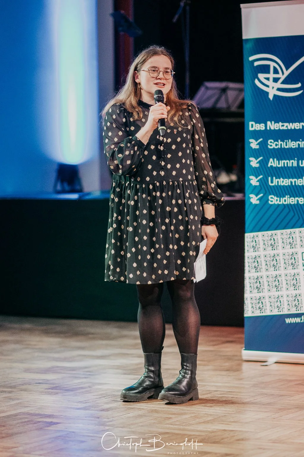 Eine junge Frau mit Brille, schwarzes Kleid mit weißen Punkten, schwarze Strumpfhosen und Stiefel, hält ein Mikrofon und spricht auf einer Bühne bei einer Veranstaltung.