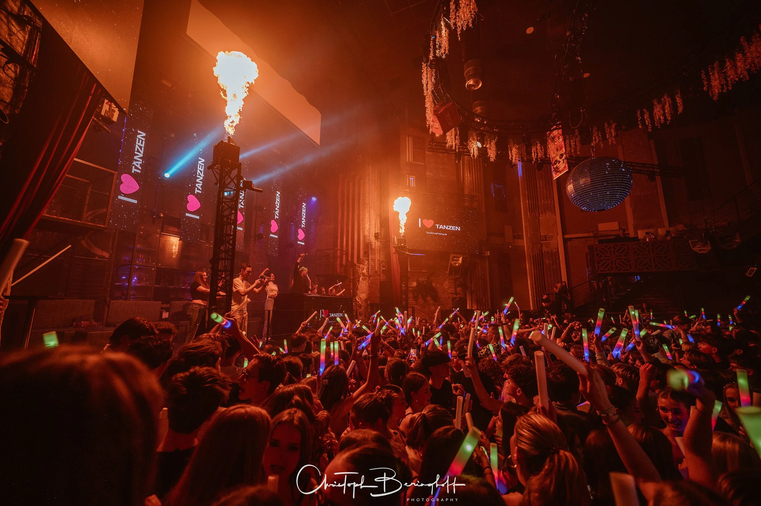 Eine große Menschenmenge tanzt in einem dunklen Club mit Lichtern und bunten Leuchtstäben, während eine Bühne mit Musikern und Flammen-Acts im Hintergrund zu sehen ist.