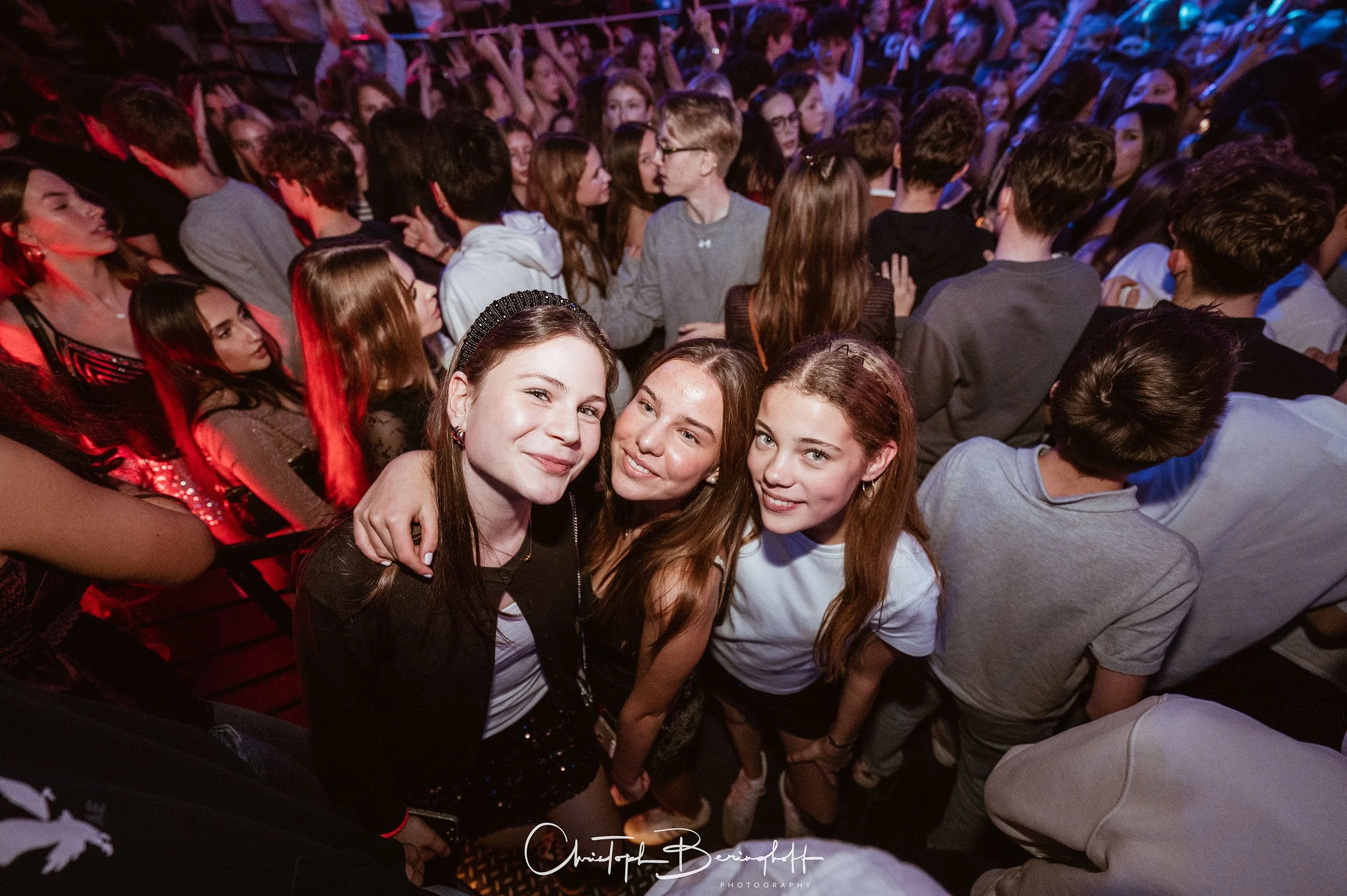 Drei junge Frauen stehen im Mittelpunkt eines großen Tanz- oder Musikfestes, umgeben von einer Menschenmenge, die im Hintergrund auf einer Party oder Veranstaltung tanzt.