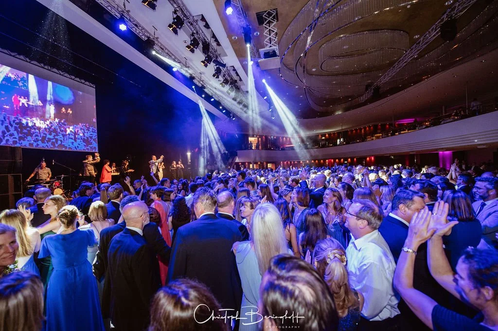 Menschenmenge bei einer Veranstaltung im großen Saal mit Bühne, Scheinwerfern und einer großen Leinwand, die eine Band auf der Bühne zeigt.