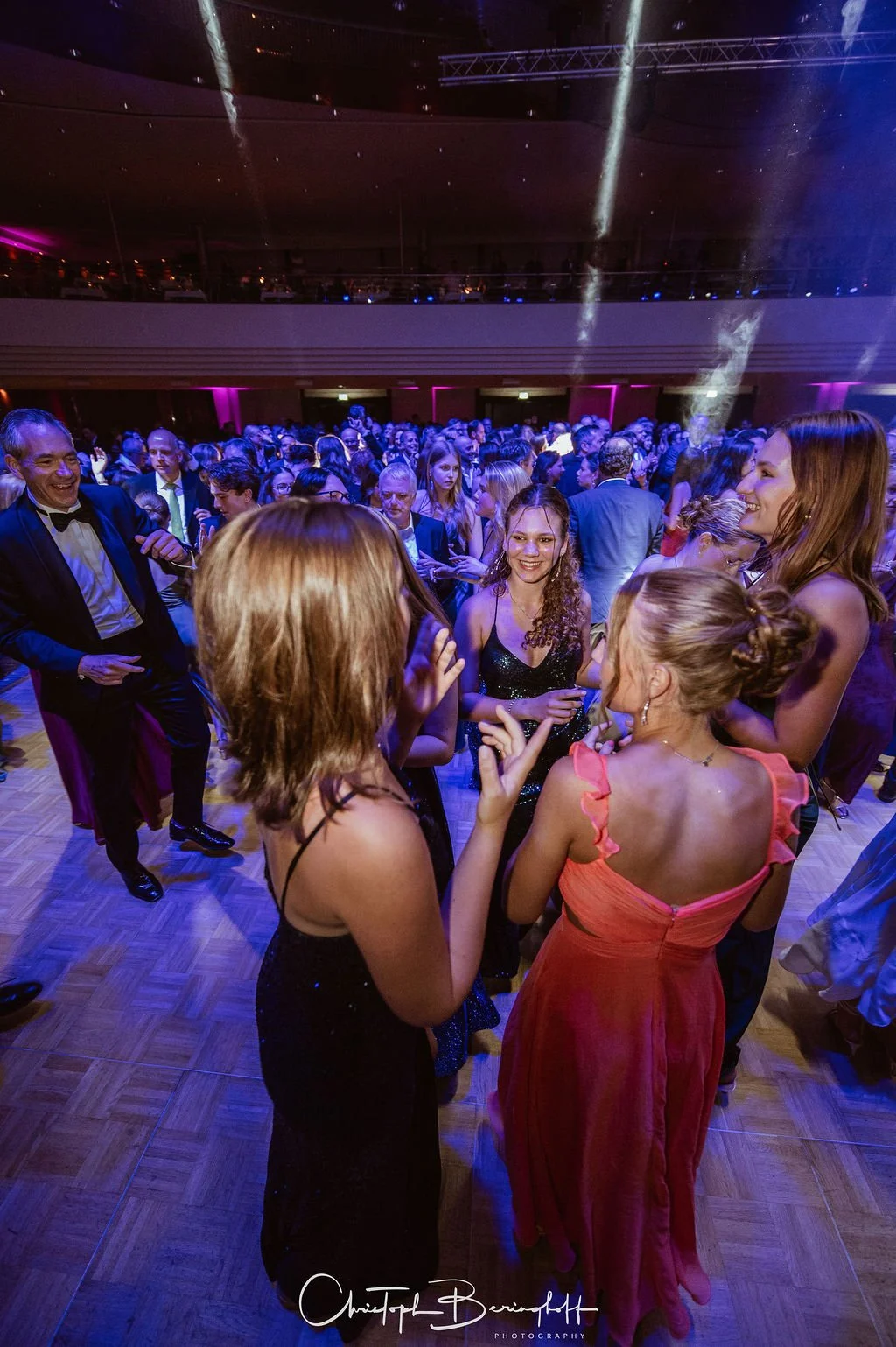 Menschen tanzen und unterhalten sich auf einer eleganten Party in einem großen Saal mit Tanzfläche, farbigen Lichtern und deutlichem Festambiente.