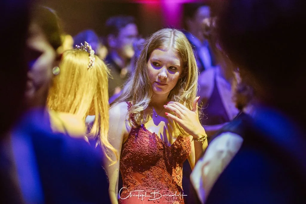 Junge Frau in rotem Spitzenkleid bei einer Party, umgeben von anderen Gästen in dunkler Umgebung mit bunter Beleuchtung.