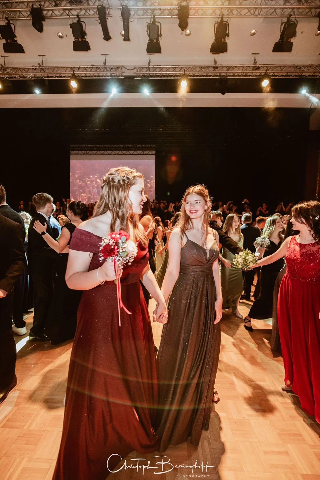 Junge Frauen tanzen auf einer Party im stilvollen Abendkleid. Im Hintergrund viele weitere Gäste und eine große Leinwand. Die Stimmung ist fröhlich.