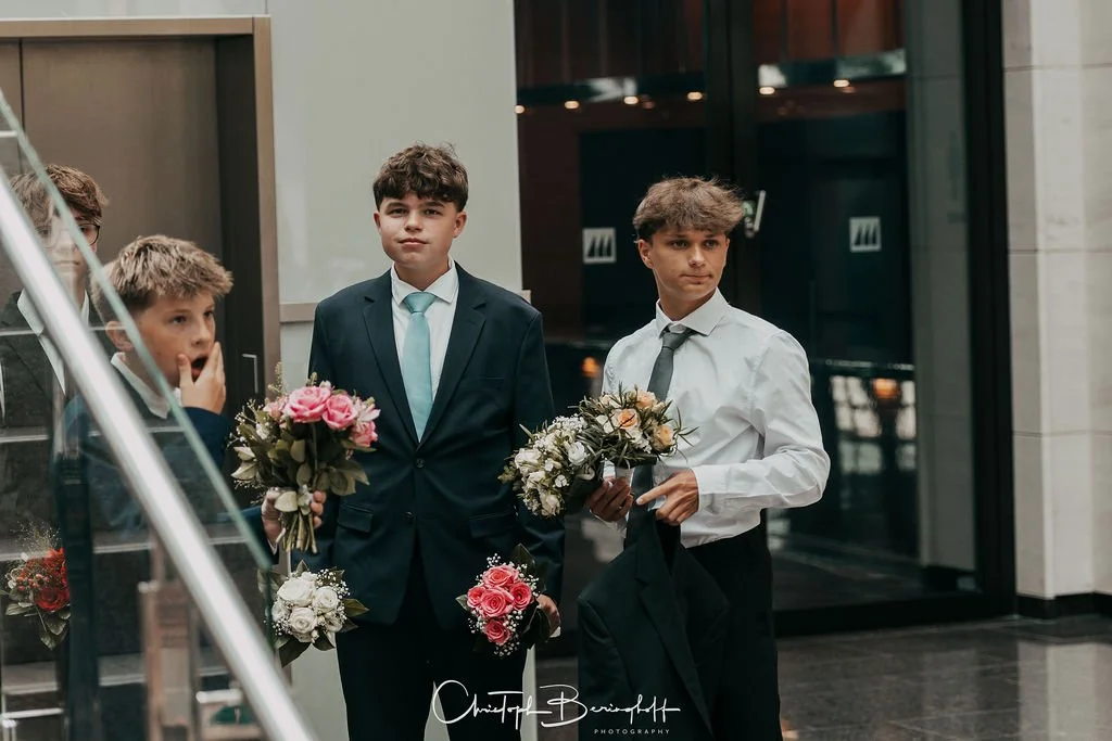 Drei Jungen in formeller Kleidung mit Blumensträußen, vermutlich bei einer Hochzeitsfeier oder einem ähnlichen Anlass, in einem modernen Innenraum.