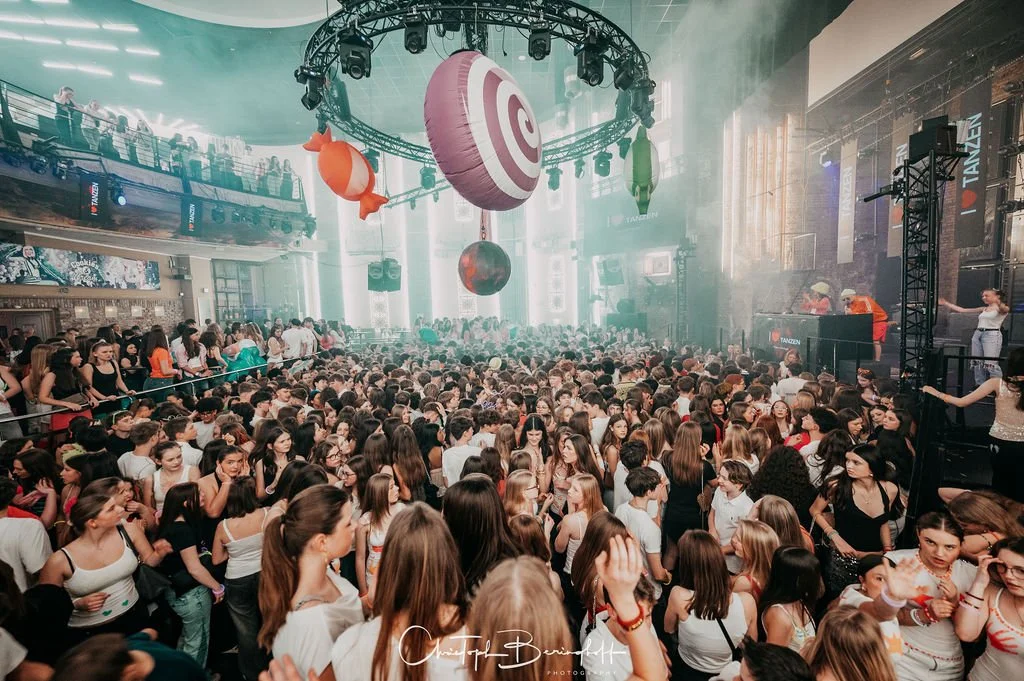 A large crowd is dancing and partying in a club with light and fog effects, a stage with DJs, and decorations such as a target, a fish, and a globe.