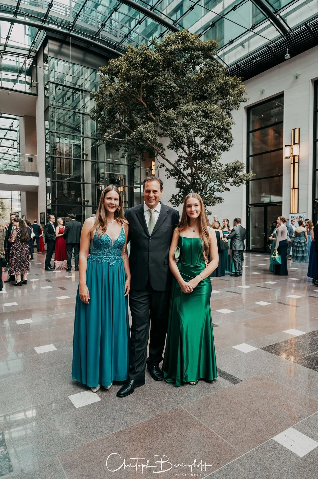Zwei junge Frauen in eleganten Kleidern und ein Mann in Anzug posieren in einem modernen, Glas-Indoor-Hochzeits- oder Eventraum.