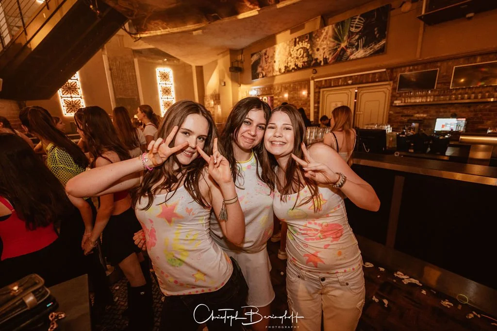 Three young women are partying in a bar, laughing and posing for the camera.