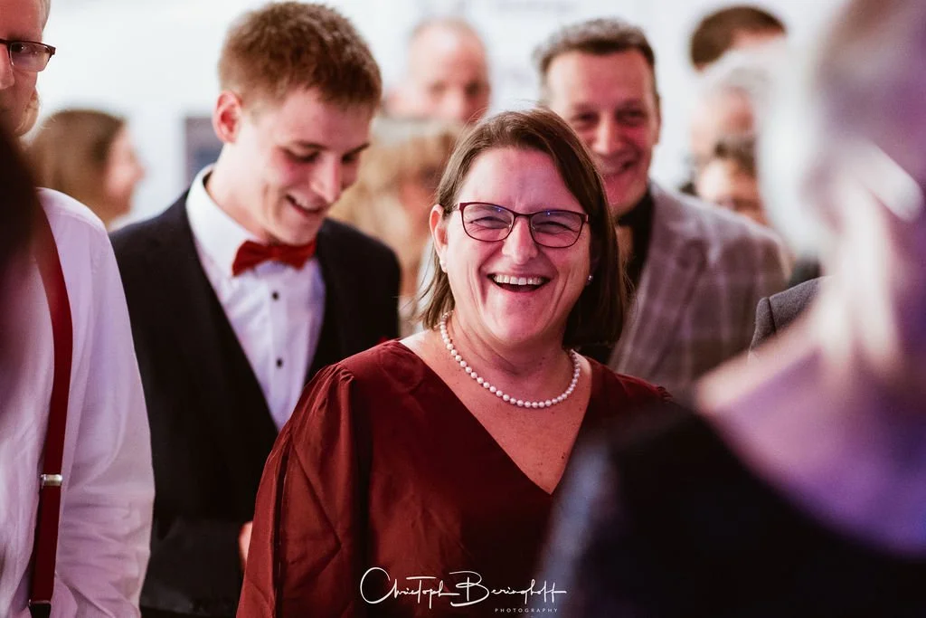 Menschen lachen und feiern bei einer Veranstaltung, zentrale Frau trägt Brille, Perlenkette, in burgunderfarbenem Kleid, umgeben von jungen und älteren Personen in eleganter Kleidung.