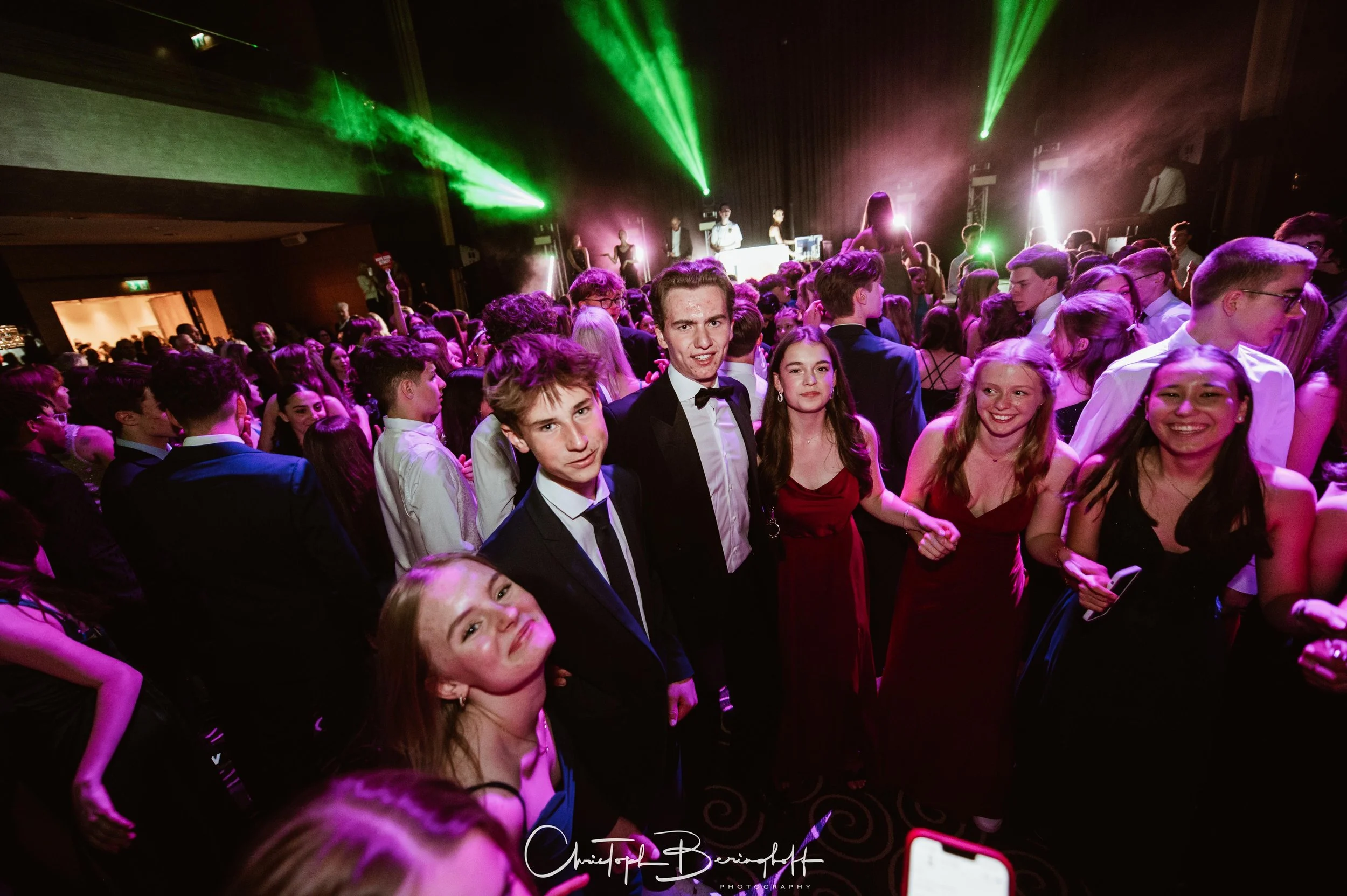 Junge Leute auf einer Gala oder Abschlussfeier, tanzend und lachend in formeller Kleidung, mit bunten Lichteffekten im Hintergrund.