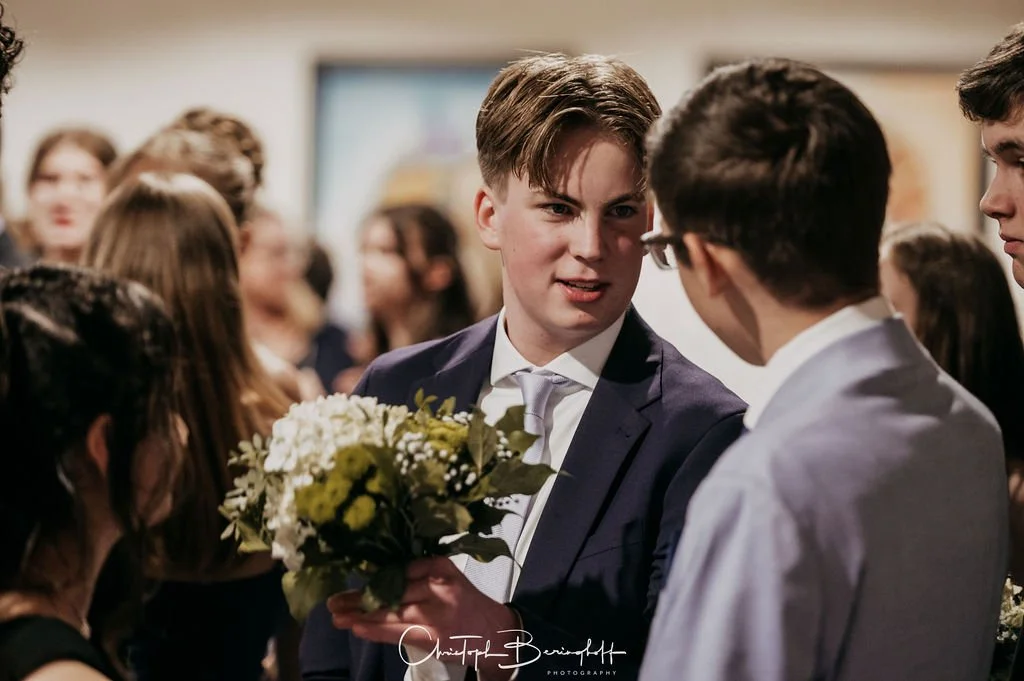 Junge Männer bei einem formellen Event, einer hält einen Blumenstrauß in der Hand.