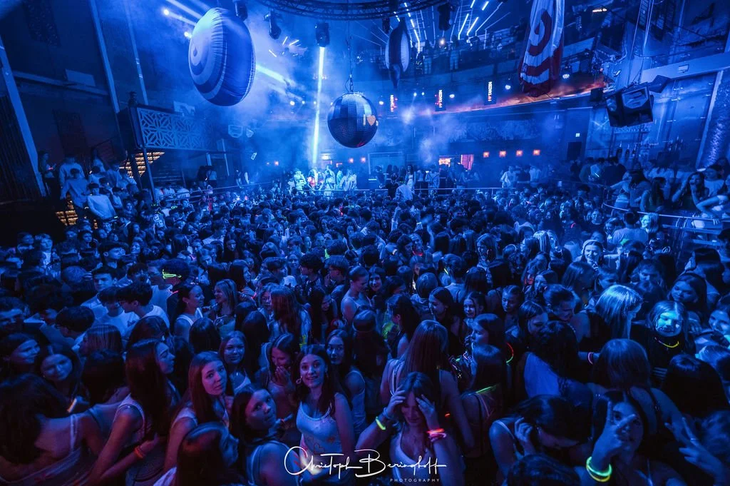 A crowd in a nightclub with blue lighting, disco balls, and a dance floor.